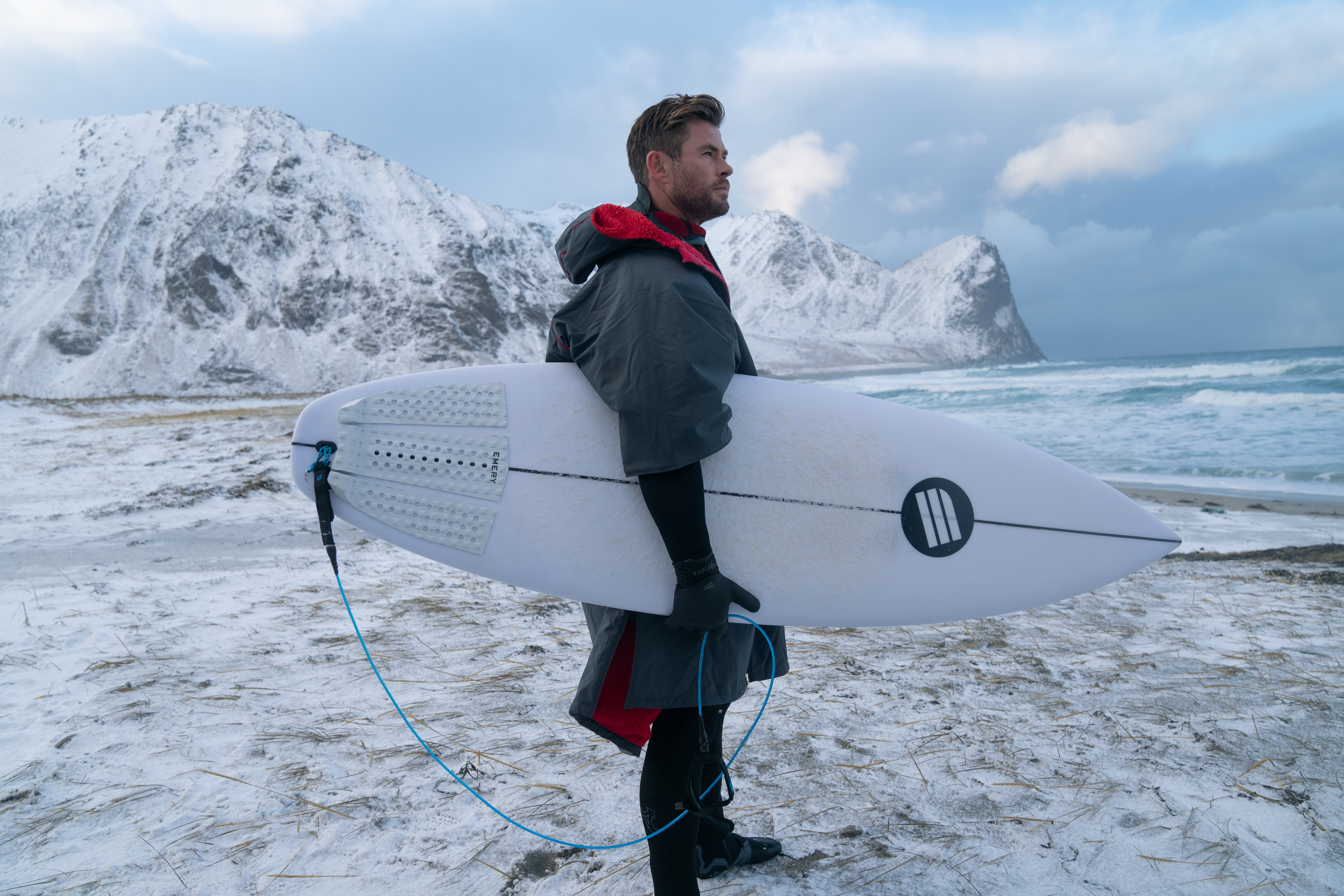 Chris Hemsworth stands on the beach with a surf board. [Photo of the day - January 2026]