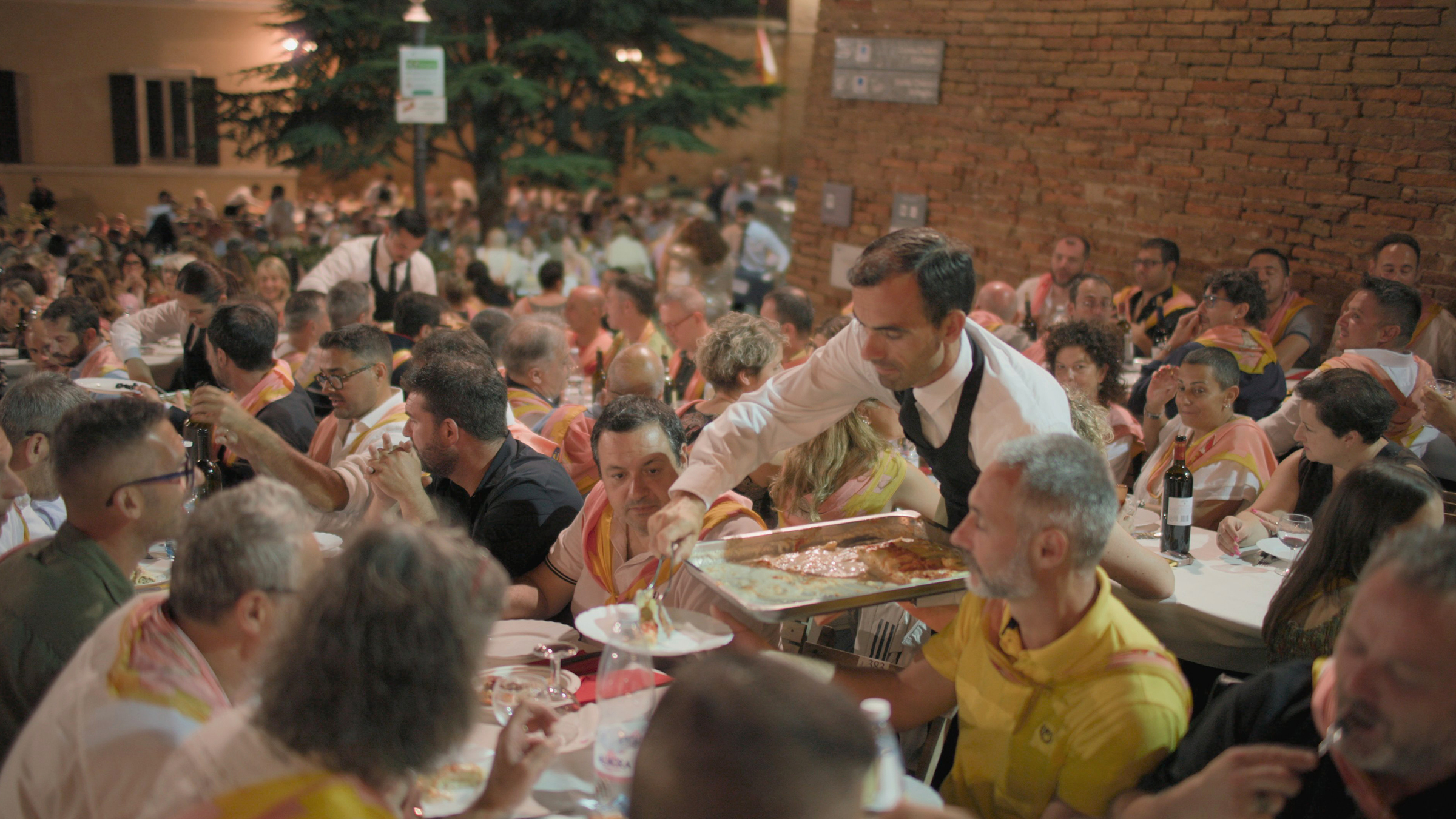 A waiter serves hot food from a tray to a participant at a bustling Palio dinner, held on the... [Photo of the day - June 2026]